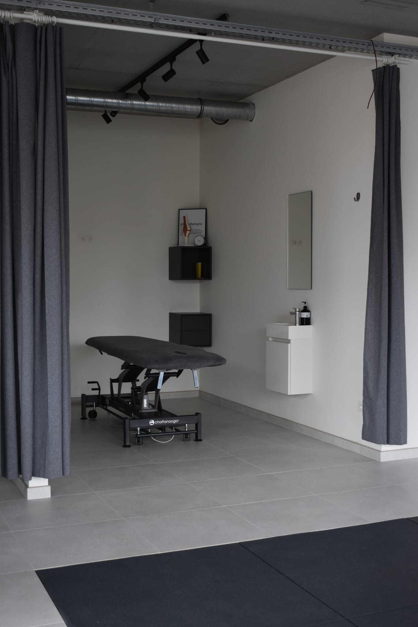 Een van de praktijkruimtes van PhysioDB, afschermbaar door een gordijn en uitgerust met een electrische kinesietherapie tafel en lavabo.